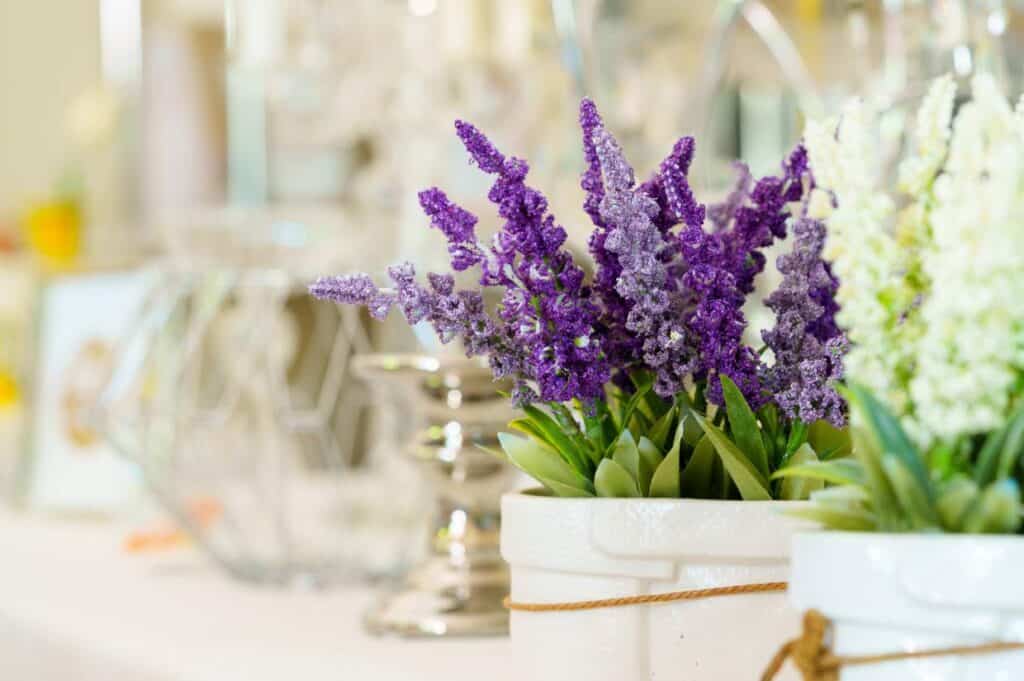 A close-up of purple flowers arranged in a white pot on a decorated table with blurred glassware in the background.