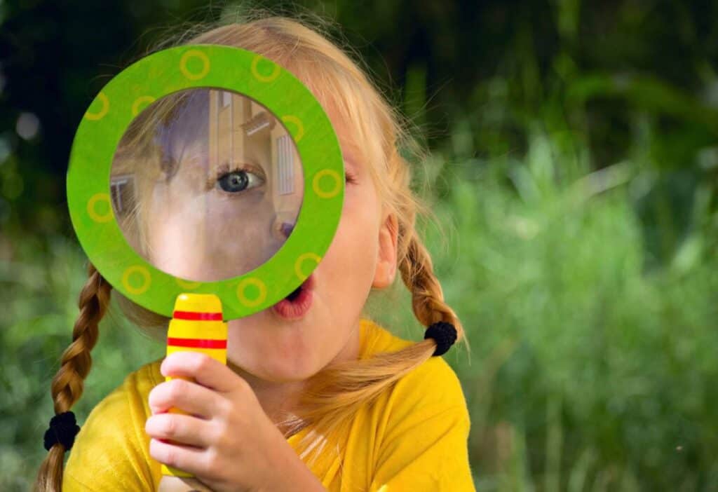 A child with blonde hair in pigtails is holding a magnifying glass up to their face, distorting one eye. The child is wearing a yellow shirt and is outdoors in a grassy area.