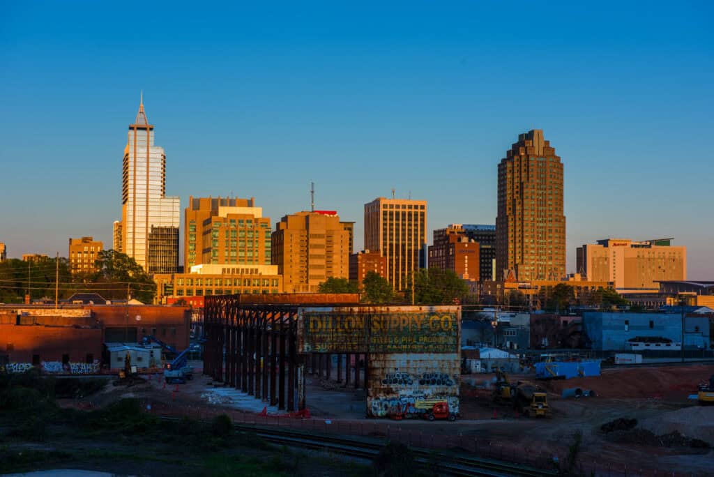 A city skyline at sunset, with a mix of modern and older buildings, an unfinished construction site in the foreground, and a train track running through the area.