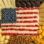 An assortment of snacks is arranged to resemble the American flag, including blueberries, strawberries, cream, wafers, cookies, pretzels, graham crackers, and chocolate pieces.