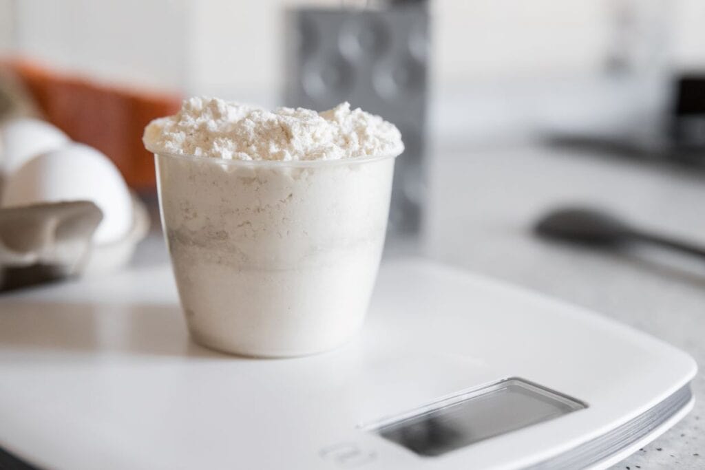A measuring cup filled with flour is placed on a digital kitchen scale. Eggs and a spoon are in the background.