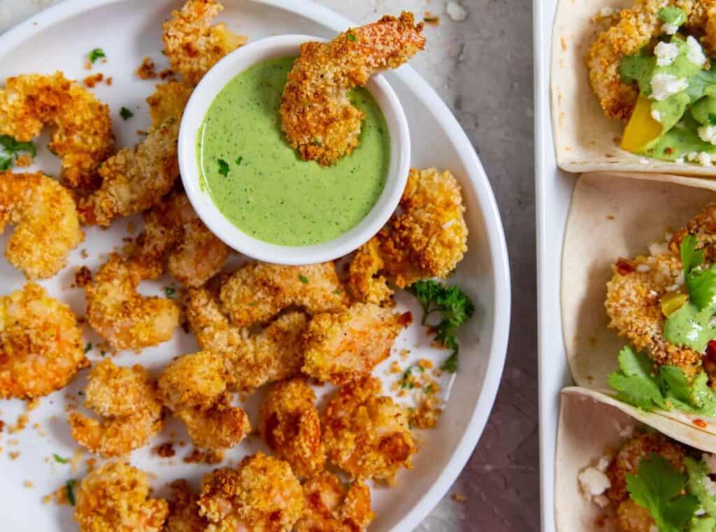 Plate of crispy fried shrimp with a green dipping sauce in a bowl at the center, and taco shells filled with various ingredients on the side.