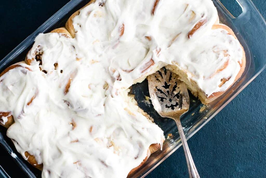 A glass baking dish containing frosted cinnamon rolls, with a portion missing and a metal serving spatula resting in the dish.