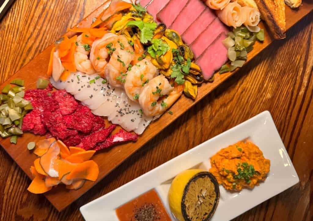 A wooden board with an assortment of sliced meats, shrimp, pickles, and vegetables, accompanied by a rectangular plate with a charred lemon, orange spread, and dipping sauce.