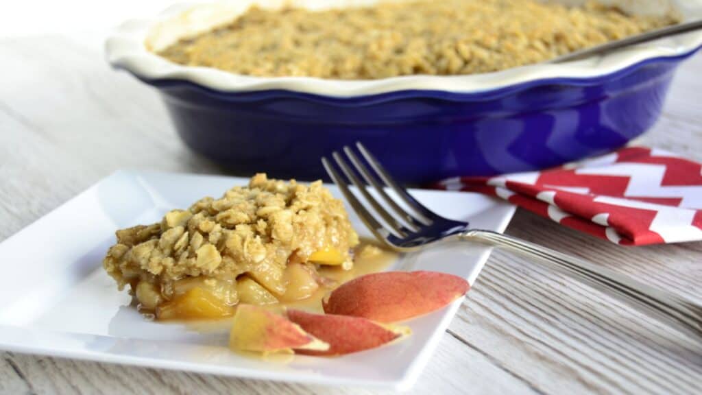 A square white plate with a serving of peach cobbler and two peach slices placed in front of a blue baking dish filled with peach cobbler. A fork and a red-and-white napkin are nearby.
