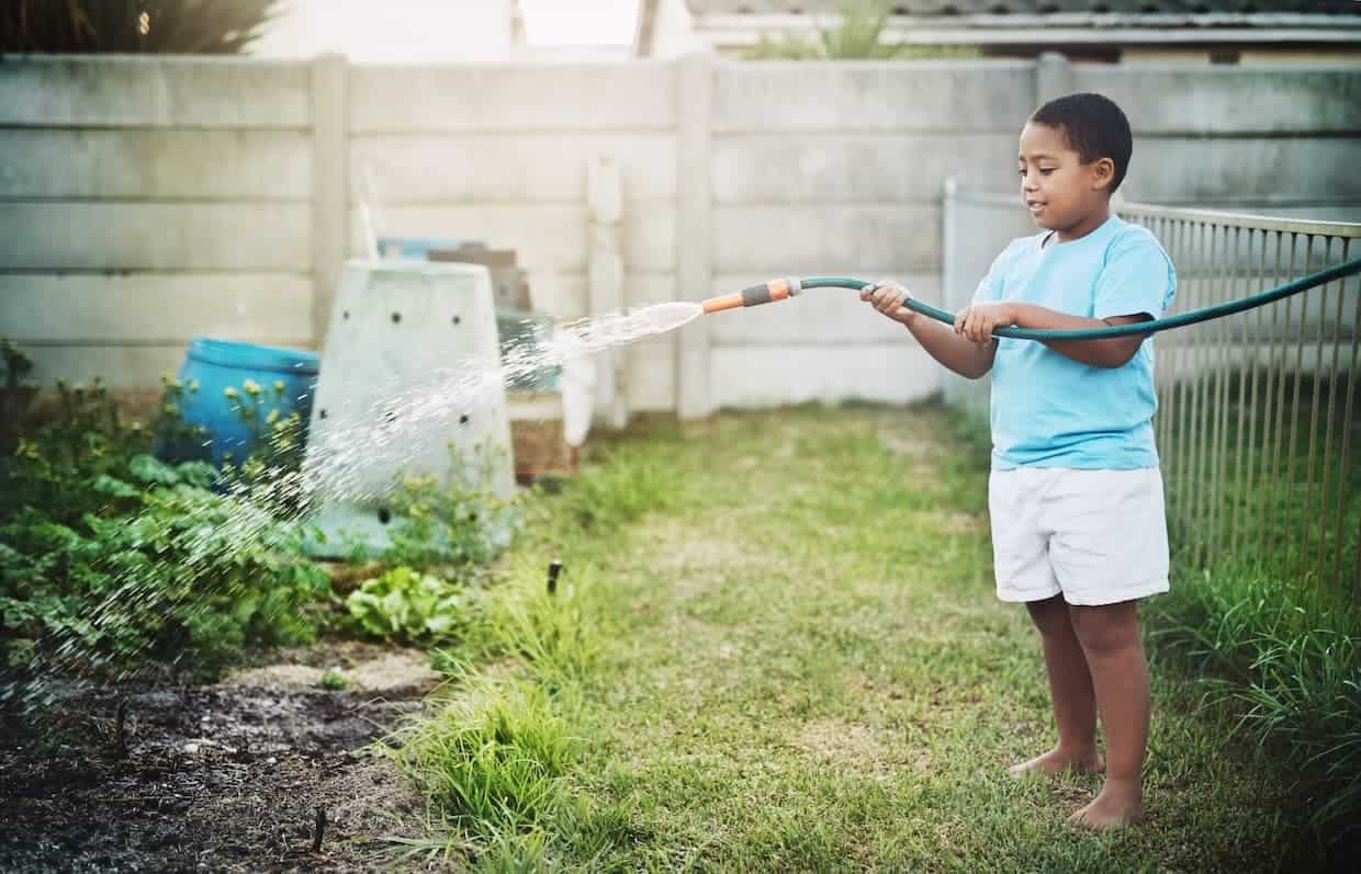 A child in a light blue shirt and white shorts waters a garden with a hose in a backyard.