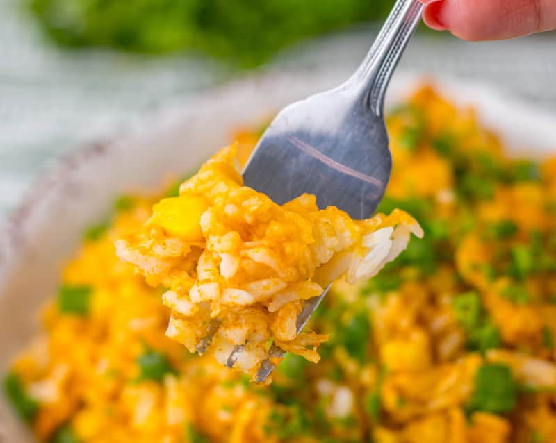 A fork holding a bite of cheesy rice dish with visible pieces of yellow corn and green herbs, with the main bowl of the dish blurred in the background.