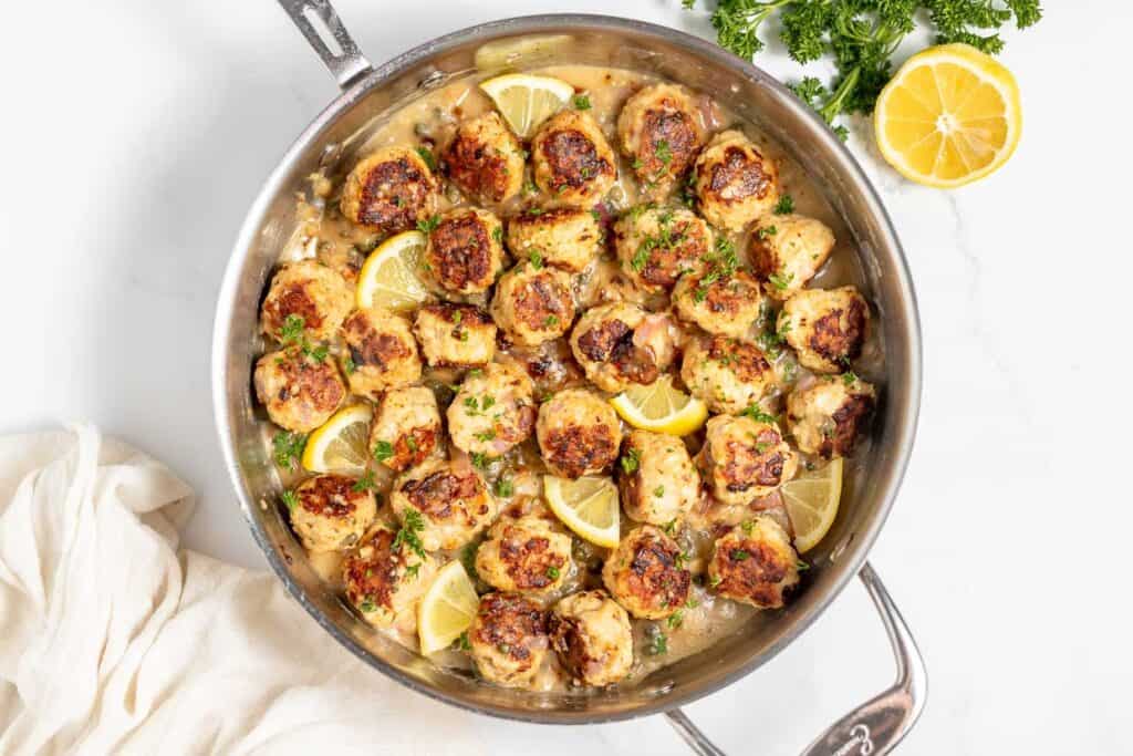 A skillet filled with browned meatballs in a creamy sauce garnished with lemon wedges and chopped parsley. A lemon half and a bunch of parsley are placed nearby on a marble surface.