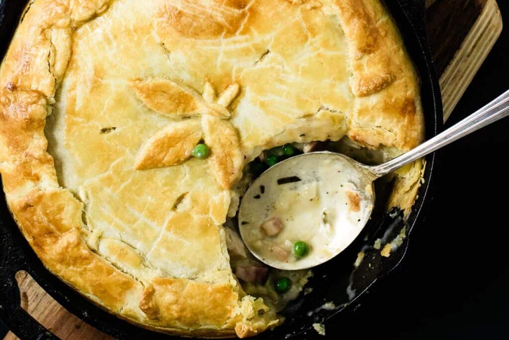A close-up of a pot pie with a golden-brown crust, partially cut to reveal a creamy filling with peas and chunks of meat. A spoon rests in the filling, indicating it has been served—one of the timeless May recipes enjoyed by many.