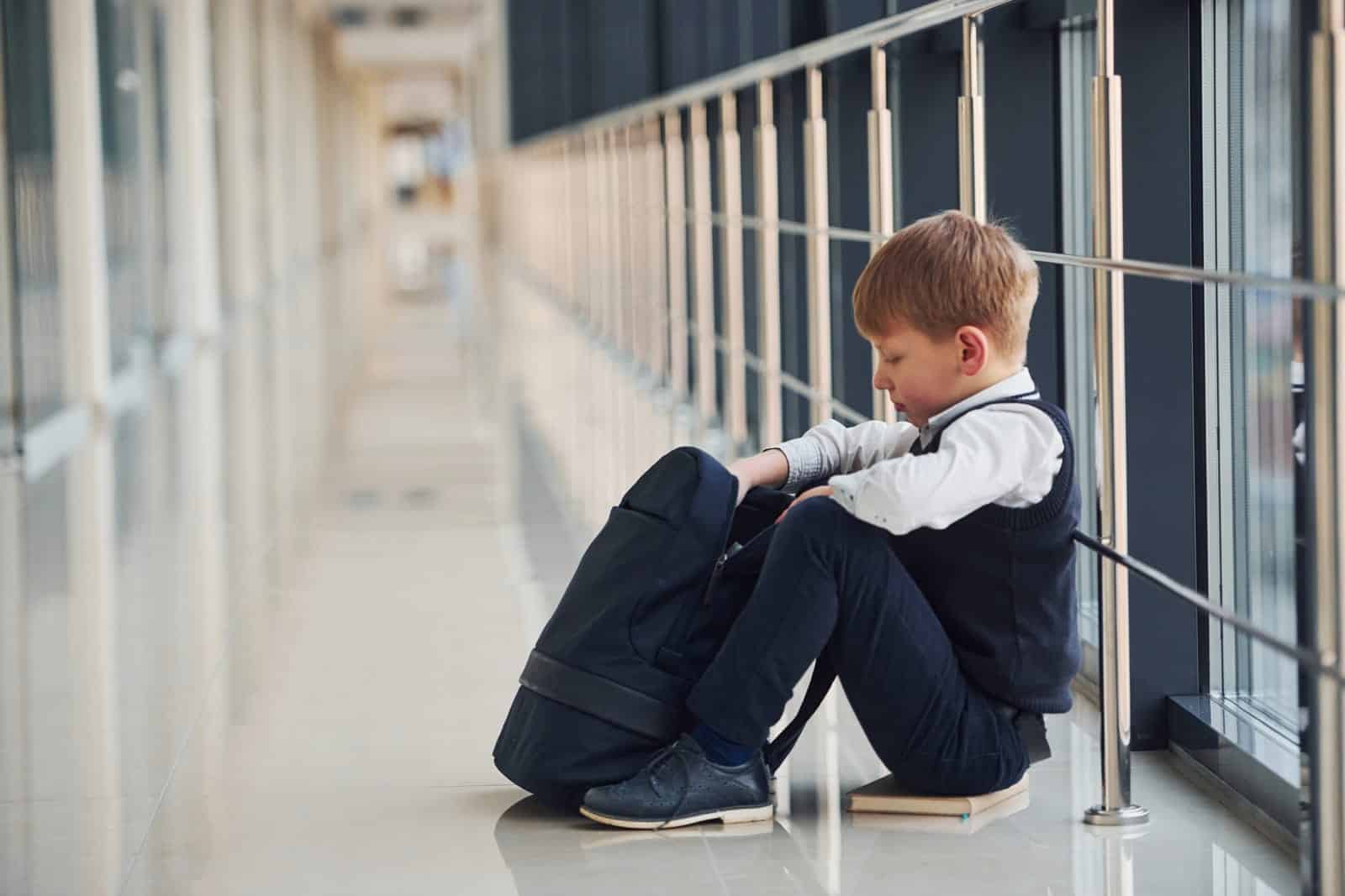 A young boy in a school uniform sits on the floor in a long hallway, looking into his black backpack.