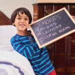 A smiling child holding a chalkboard with a list of chores. "Dishes" and "Laundry" are checked off, while "Clean Room" is not. The child stands next to a bed and a wooden dresser.