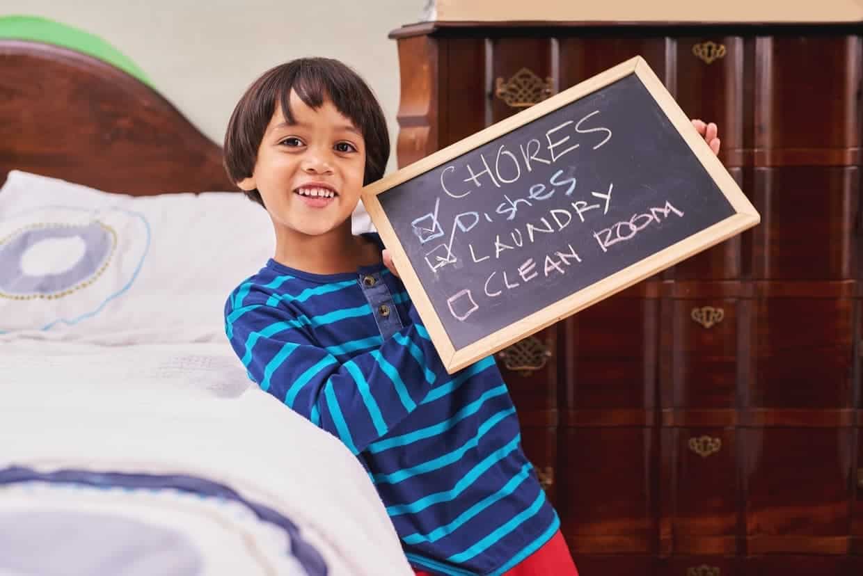 A smiling child holding a chalkboard with a list of chores. "Dishes" and "Laundry" are checked off, while "Clean Room" is not. The child stands next to a bed and a wooden dresser.
