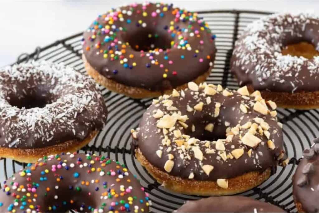 A wire rack with six chocolate-glazed donuts. Toppings include colorful sprinkles, crushed nuts, and shredded coconut.