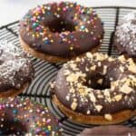 A wire rack with six chocolate-glazed donuts. Toppings include colorful sprinkles, crushed nuts, and shredded coconut.