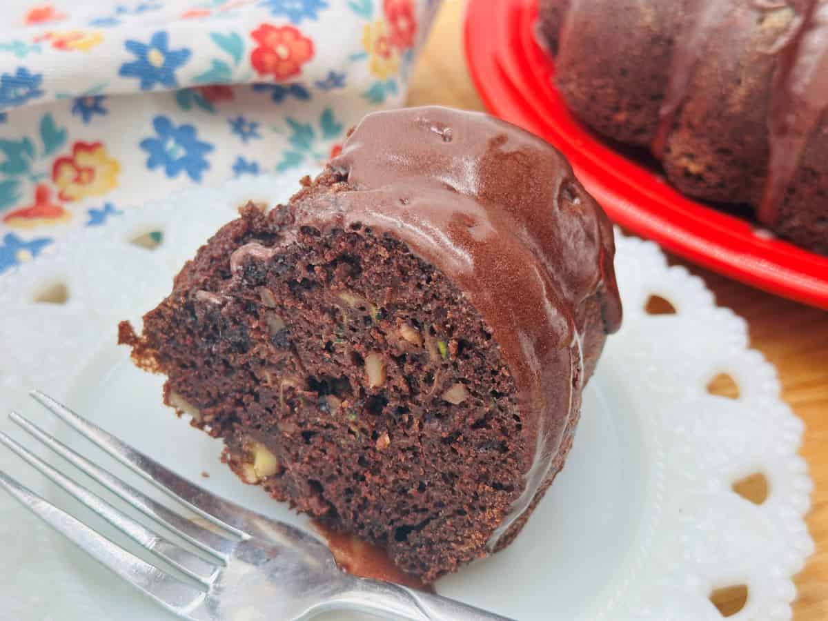 A slice of chocolate bundt cake with walnuts on a white plate, accompanied by a fork. The cake is partially seen on a red plate in the background, with a floral napkin next to it.