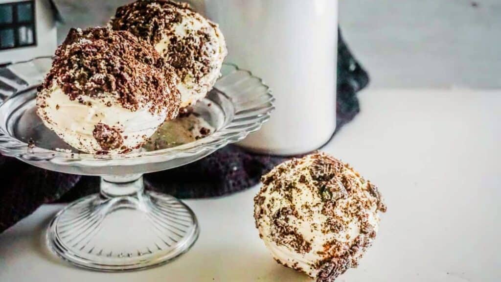 A glass dessert stand holds two cookies and cream hot chocolate bombs.