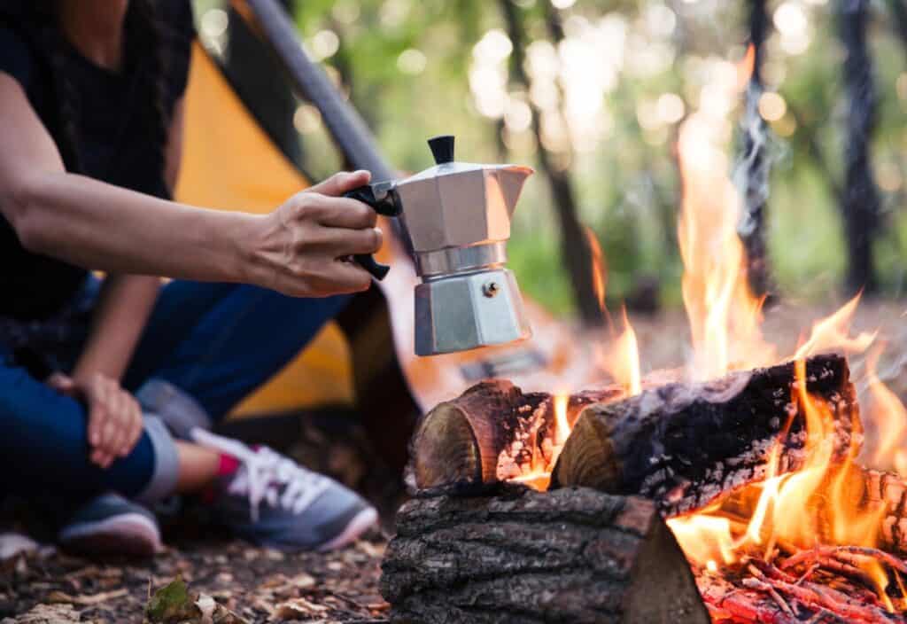 A person holds a moka pot over a campfire with a yellow tent visible in the background, surrounded by trees.