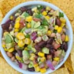 A bowl of bean and corn salad with diced avocado, red onion, tomatoes, and cilantro surrounded by tortilla chips.