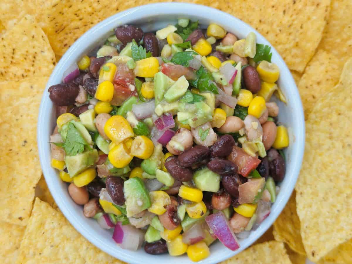 A bowl of bean and corn salad with diced avocado, red onion, tomatoes, and cilantro surrounded by tortilla chips.