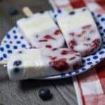 Three berry yogurt popsicles on a white plate with blue polka dots, placed next to a red and white checkered cloth.