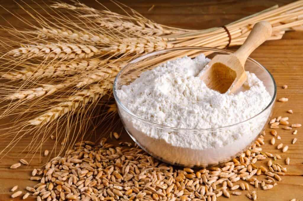 A bowl of flour with a wooden scoop sits on a wooden surface. Surrounding it are scattered wheat grains and a bundle of wheat stalks.