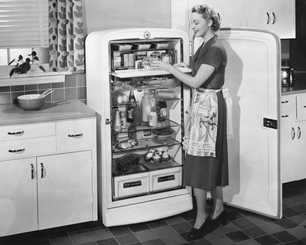 A woman in a dress and apron stands in a kitchen, organizing food in an open refrigerator. The kitchen has retro-style cabinets and checkered tile flooring.