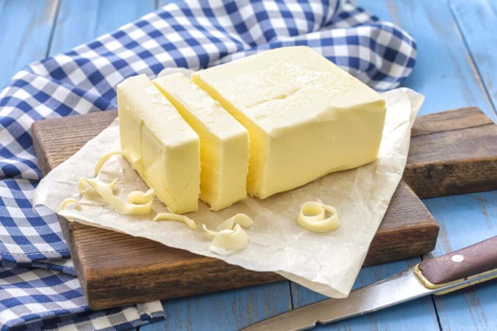 A block of butter with three slices cut off, on a piece of parchment paper atop a wooden board. A blue and white checkered cloth is in the background, and a butter knife rests nearby—perfect for a touch of elegance in dairy-free cooking.