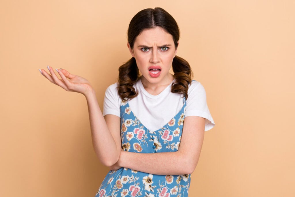 A woman with brown hair in pigtails, wearing a floral dress over a white shirt, stands against a beige background with an expression of confusion or frustration, gesturing with her right hand.