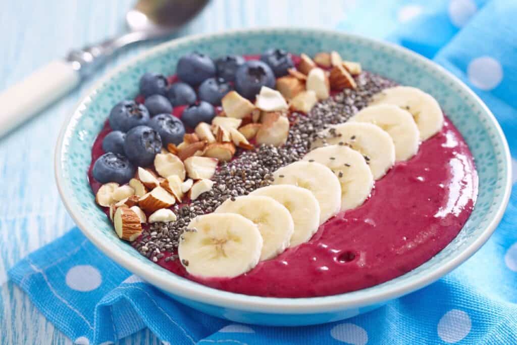 A smoothie bowl topped with sliced bananas, chia seeds, chopped nuts, and blueberries on a blue polka dot napkin with a spoon in the background highlights the joys of dairy-free cooking.