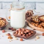A variety of nuts, including walnuts, hazelnuts, almonds, cashews, and pine nuts, are arranged around a jar of milk on a wooden surface—perfect for those exploring dairy-free cooking options.
