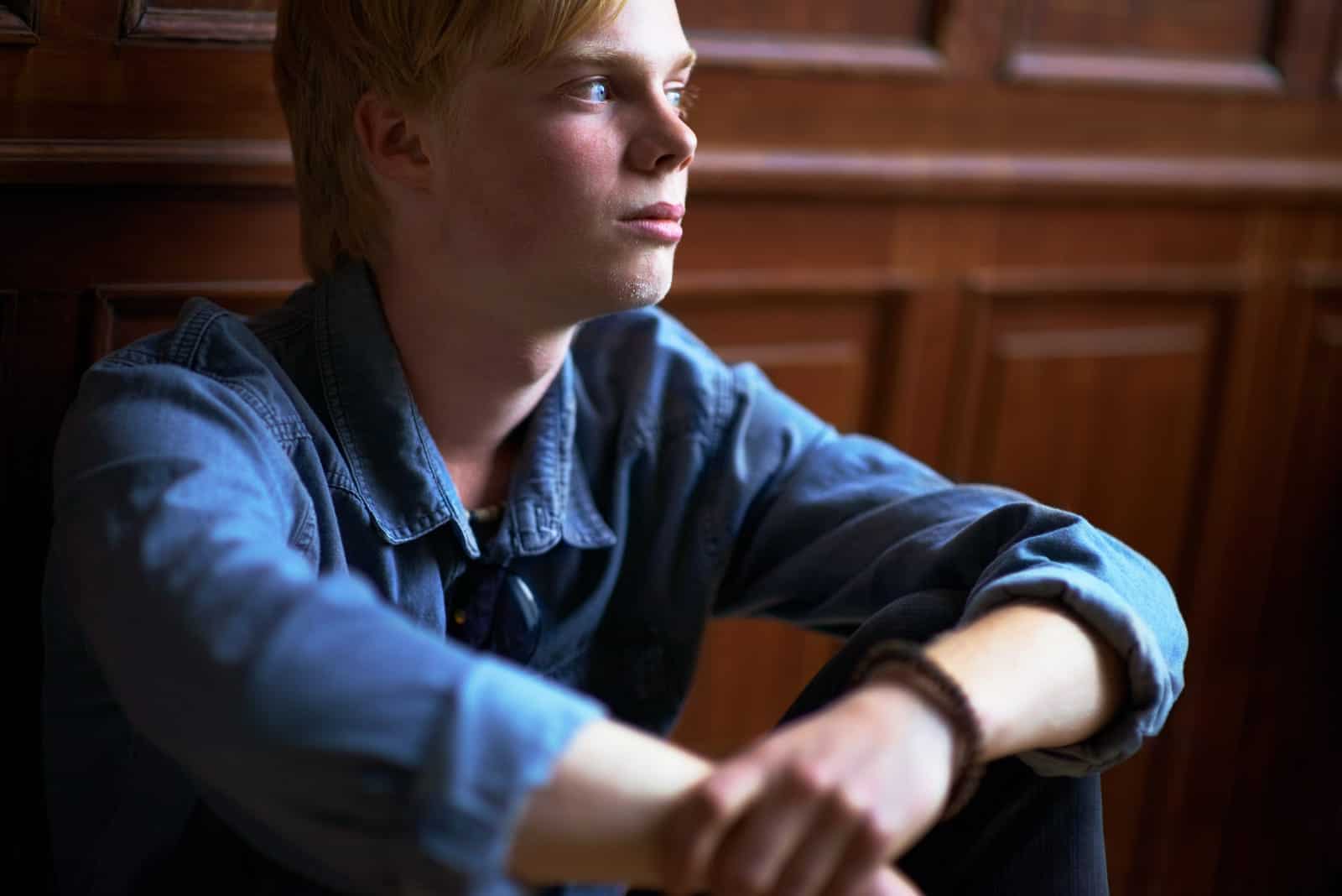 A young person in a denim jacket sits with their arms resting on their knees, gazing thoughtfully to the side. Wood paneling is visible in the background.