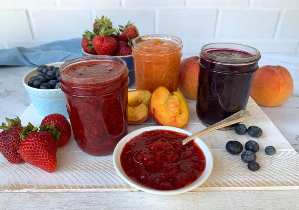 A white board displays jars of strawberry, peach, and blueberry jams, surrounded by fresh strawberries, peaches, and blueberries. Spoonful of strawberry jam in a dish is in the foreground.