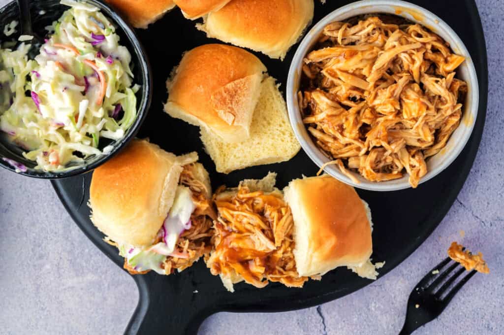 A plate with pulled chicken sliders, a bowl of coleslaw, and a fork on a light purple surface.