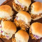 A wooden plate holds six sliders filled with shredded chicken and coleslaw, arranged neatly on a surface.