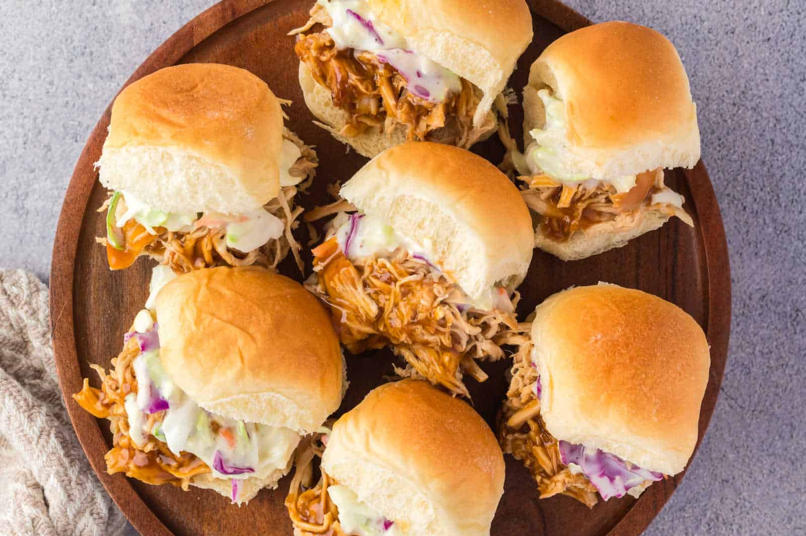 A wooden plate holds six sliders filled with shredded chicken and coleslaw, arranged neatly on a surface.