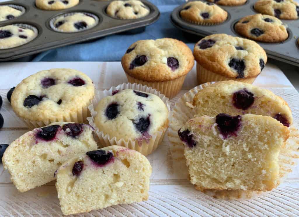 Blueberry muffins, some whole and some sliced, are displayed on a wooden surface with more muffins in trays in the background.