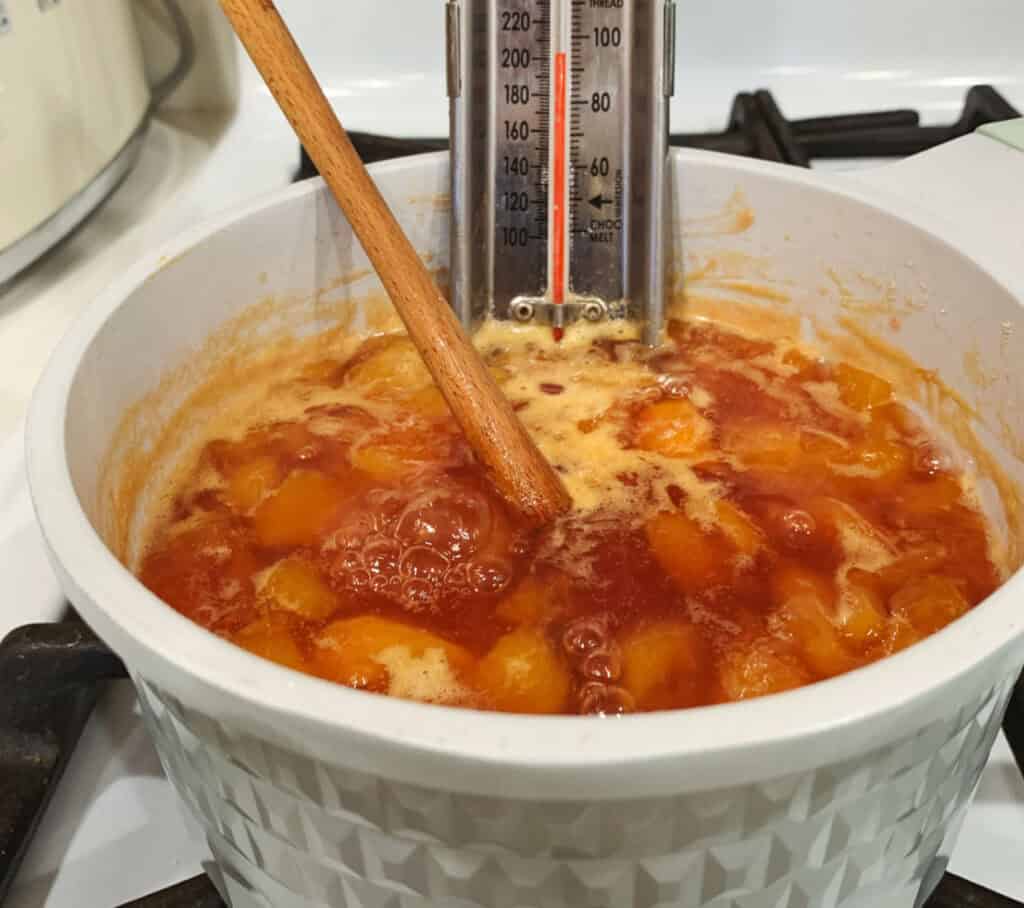 A wooden spoon stirs bubbling peach jam in a white pot on a stove. A thermometer is clipped to the pot's side, measuring the temperature of the cooking jam.