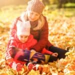 An adult and a child are sitting outdoors on fallen autumn leaves, reading a book together. The adult wears a knitted hat and jacket, and the child is dressed in a pink hat and coat.