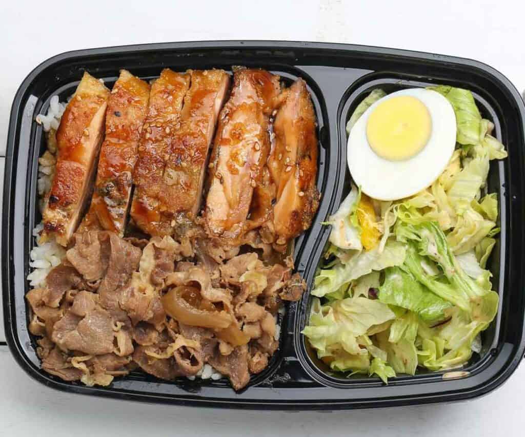 A divided food container with grilled chicken, sliced beef with onions on rice, and a side salad with iceberg lettuce and a halved boiled egg.