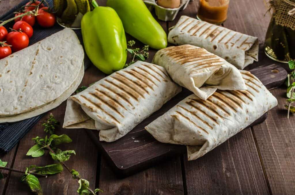 Grilled wraps on a wooden board surrounded by tortillas, fresh tomatoes, green peppers, and herbs on a wooden table.