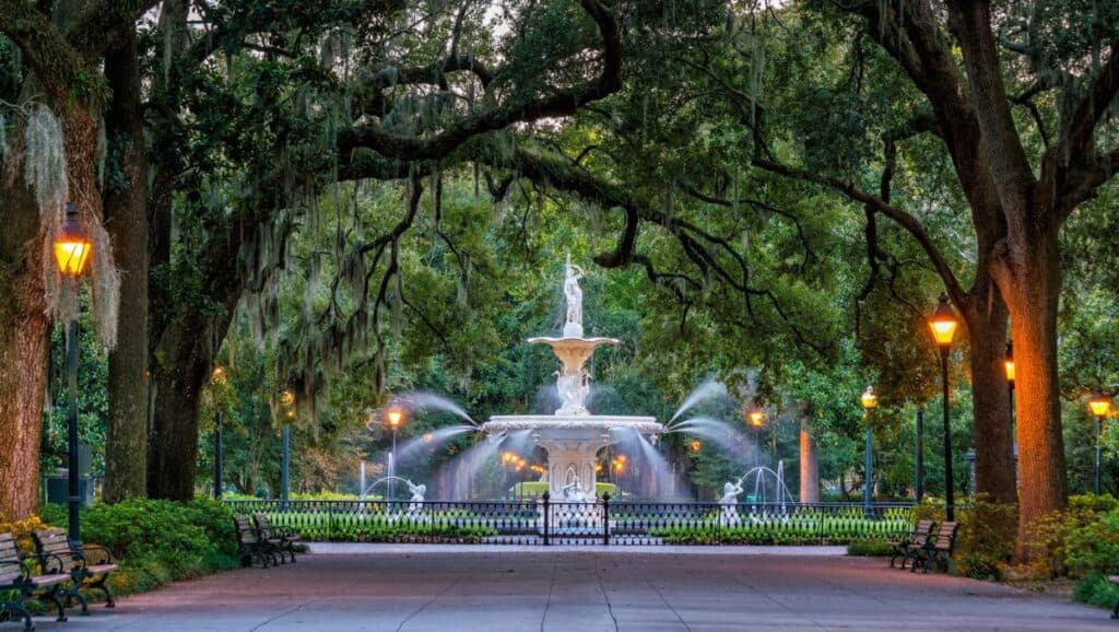 Central fountain in a park surrounded by large trees and illuminated by street lamps at dusk, with benches lining the walkway – a perfect spot to unwind among many things to do in Savannah with kids.