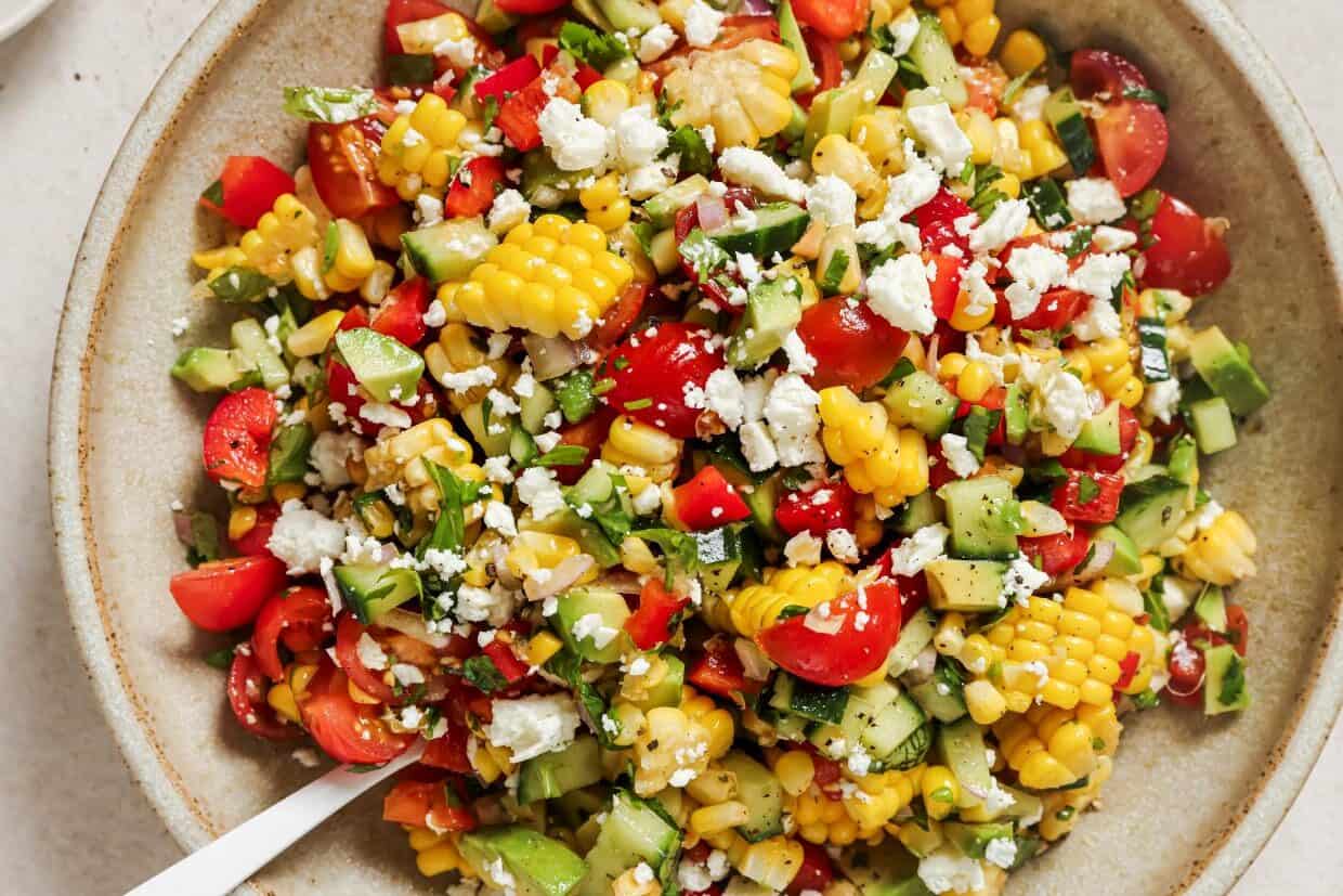 A bowl of fresh salad featuring corn, avocado, cucumber, tomatoes, and crumbled cheese, mixed with herbs.