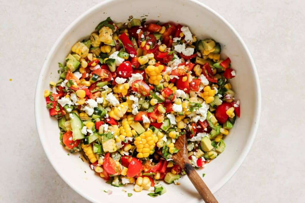 A large white bowl filled with a colorful corn salad, featuring diced avocado, tomatoes, red bell peppers, cucumbers, red onion, crumbled feta cheese, and chopped herbs, with a wooden spoon.