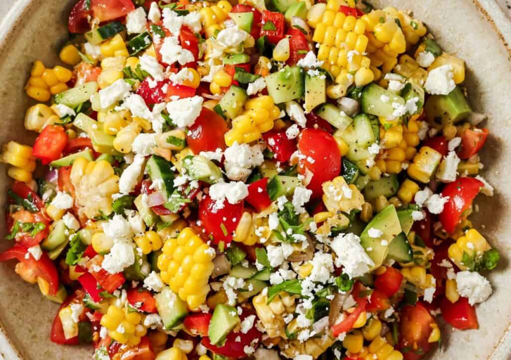 A colorful salad featuring corn, cucumbers, tomatoes, red peppers, and topped with crumbled cheese.
