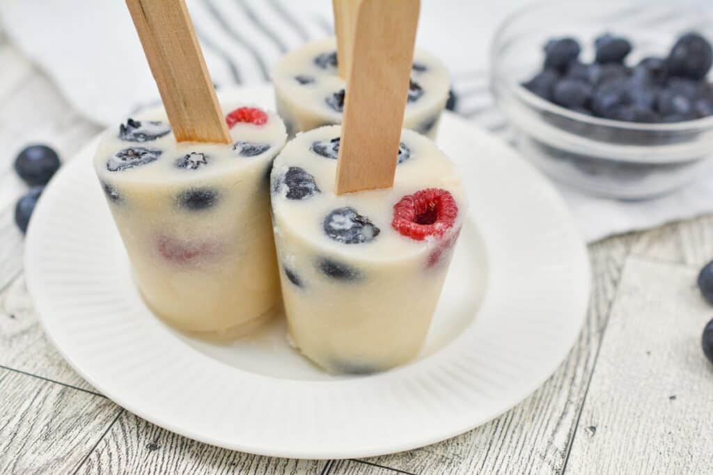 Three frozen yogurt popsicles with blueberries and raspberries served on a white plate. A bowl of blueberries is in the background.