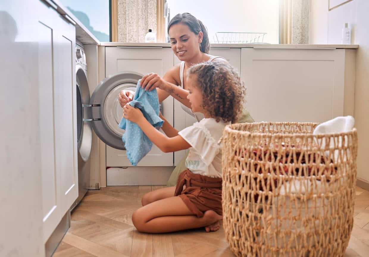 A woman and a child kneel on the floor in front of a washing machine, placing clothes inside. A laundry basket sits nearby.