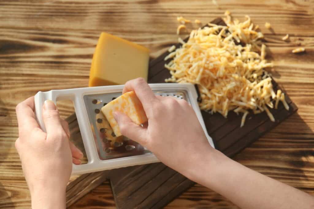 Hands grating a block of cheese on a grater, with a pile of shredded cheese and another block on a wooden cutting board in the background.