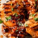 Close-up of grilled chicken pieces garnished with chopped herbs and drizzled with a glossy sauce on a light-colored plate.