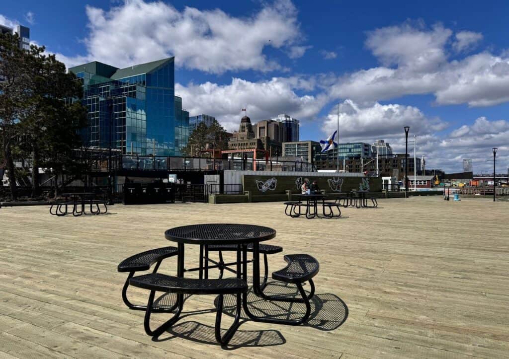 Empty circular picnic table with three benches on a spacious wooden deck, modern buildings and blue sky with clouds in the background.