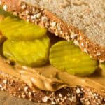 A close-up of a sandwich made with whole grain bread, featuring layers of peanut butter and sliced pickles.